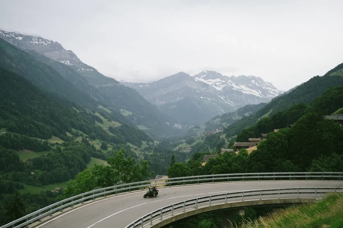 A motorcyclist rounding a curved mountain road above a deep valley with scattered settlements.