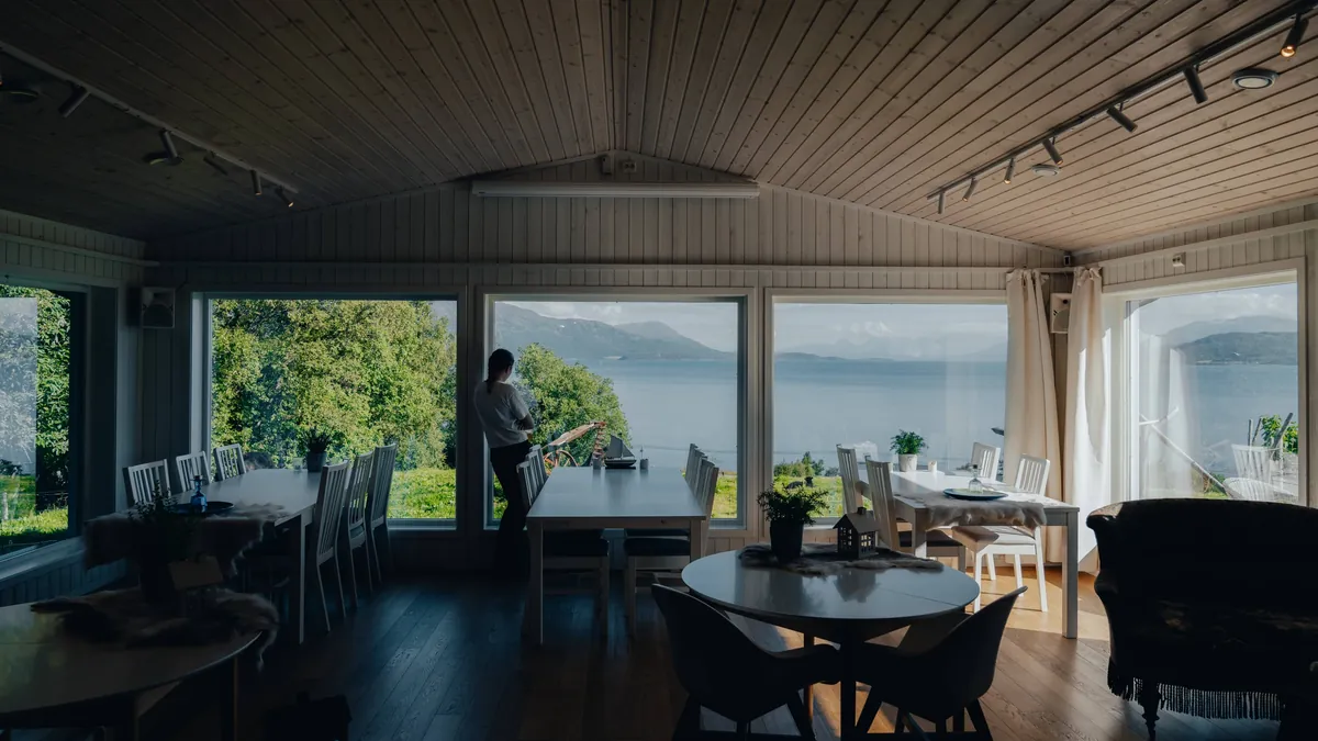 A quiet café interior with tables and chairs, and a person beside a window overlooking water.