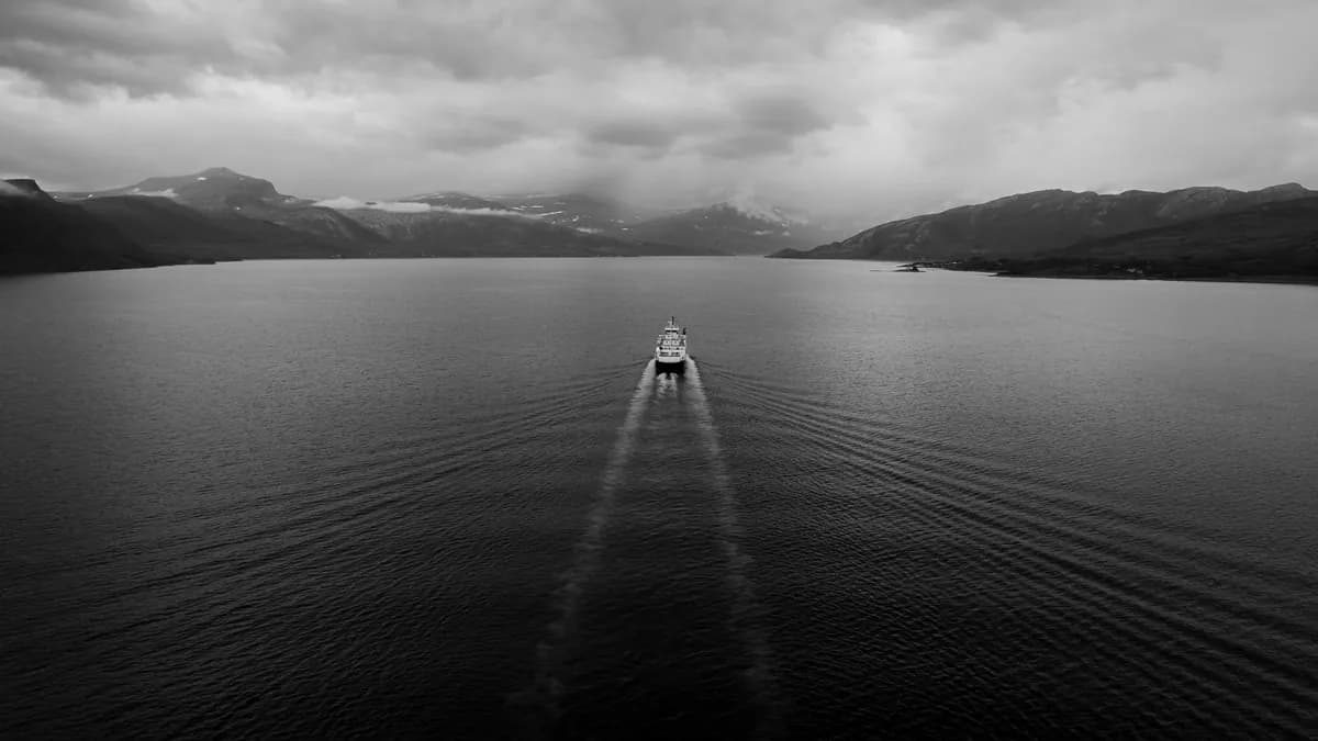 An overhead black-and-white view of a ferry and its widening wake between dark mountains.