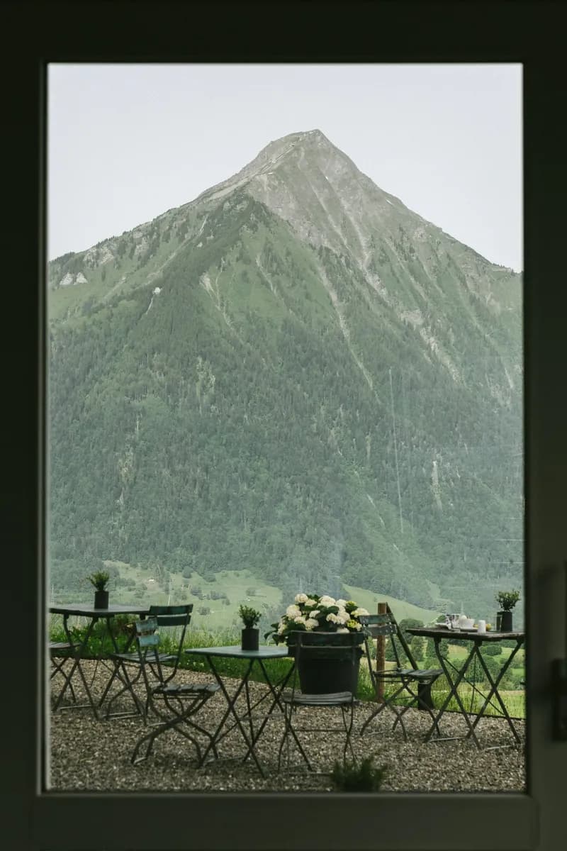 A dark window frame enclosing a mountain peak and an empty terrace with chairs.