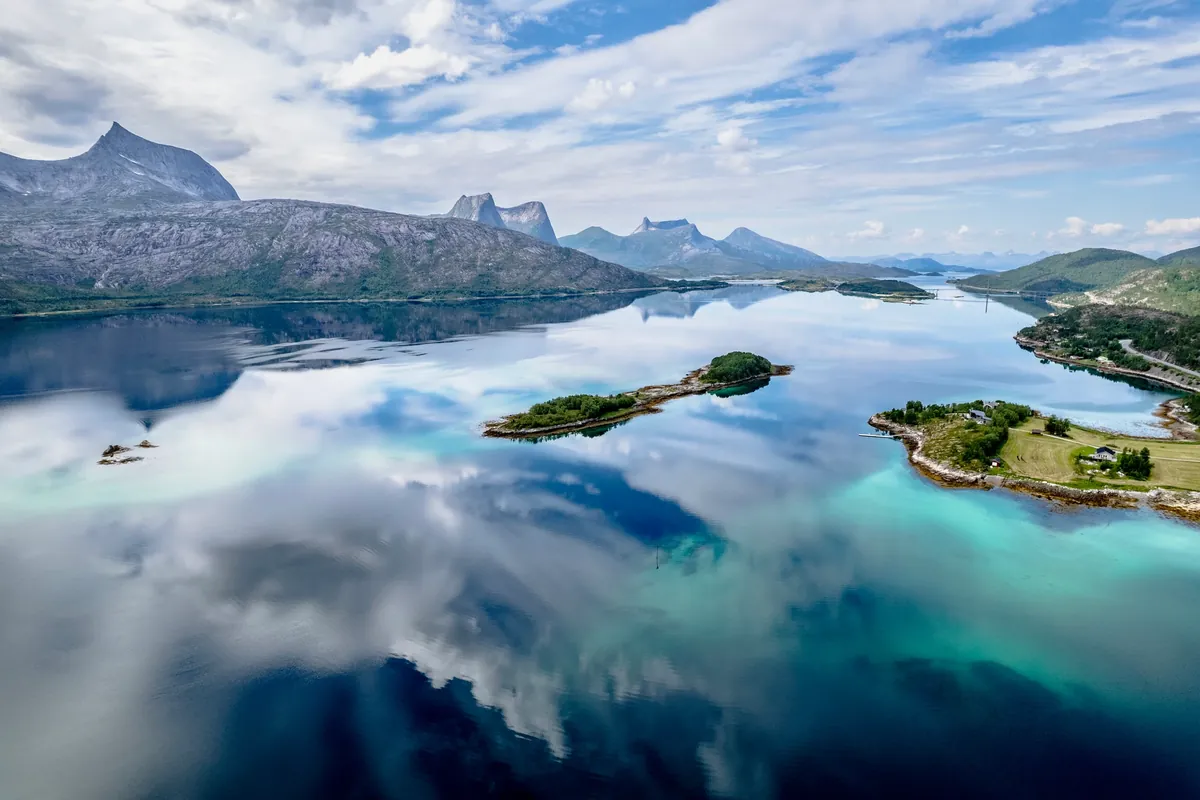 Small green islands in clear water reflecting mountains and clouds.