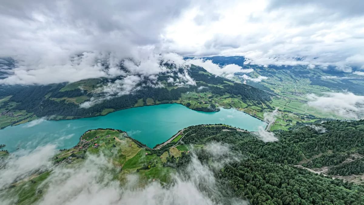 A turquoise lake surrounded by forested slopes and drifting patches of low cloud.