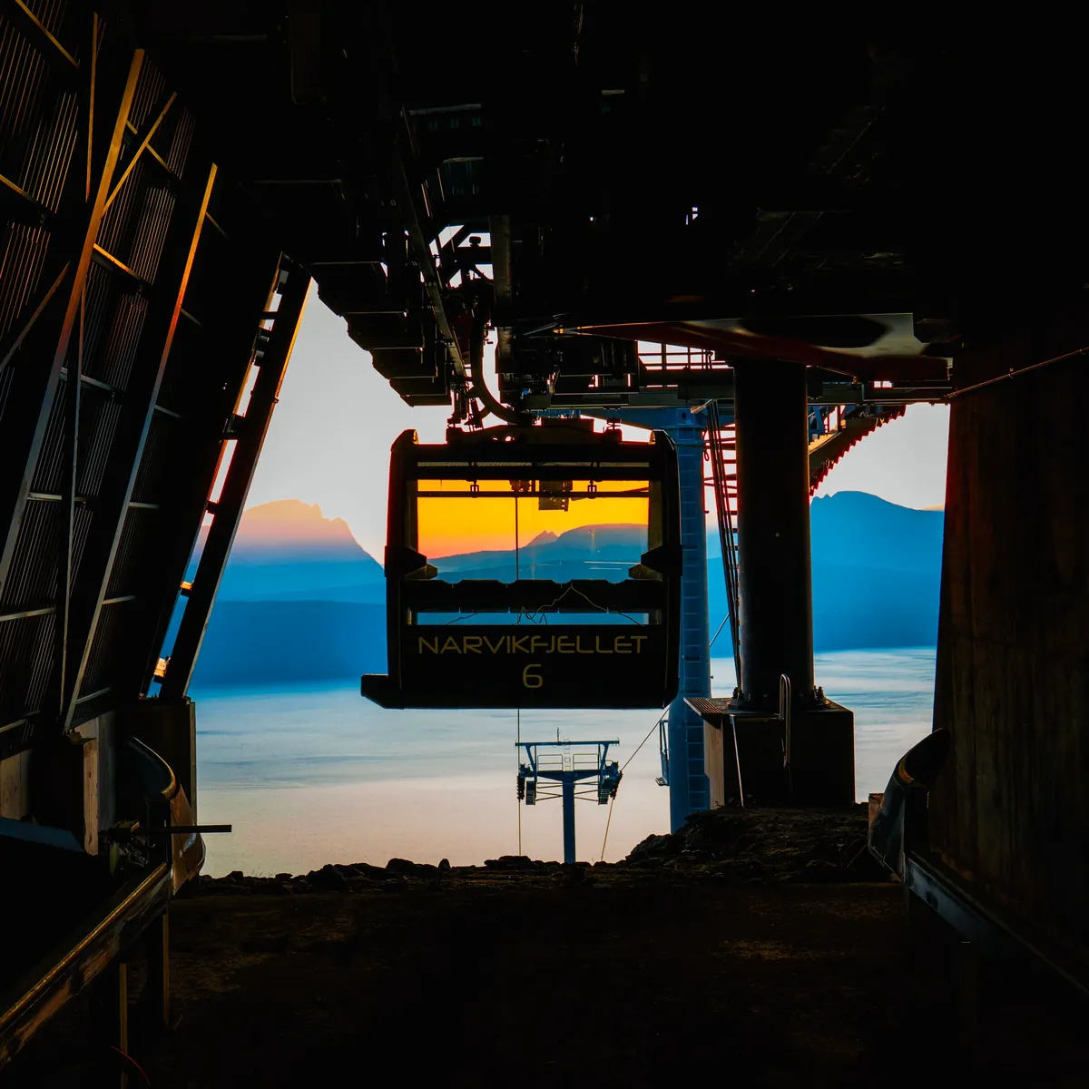 A cable car framed by dark station machinery, with an orange horizon and blue mountains beyond.
