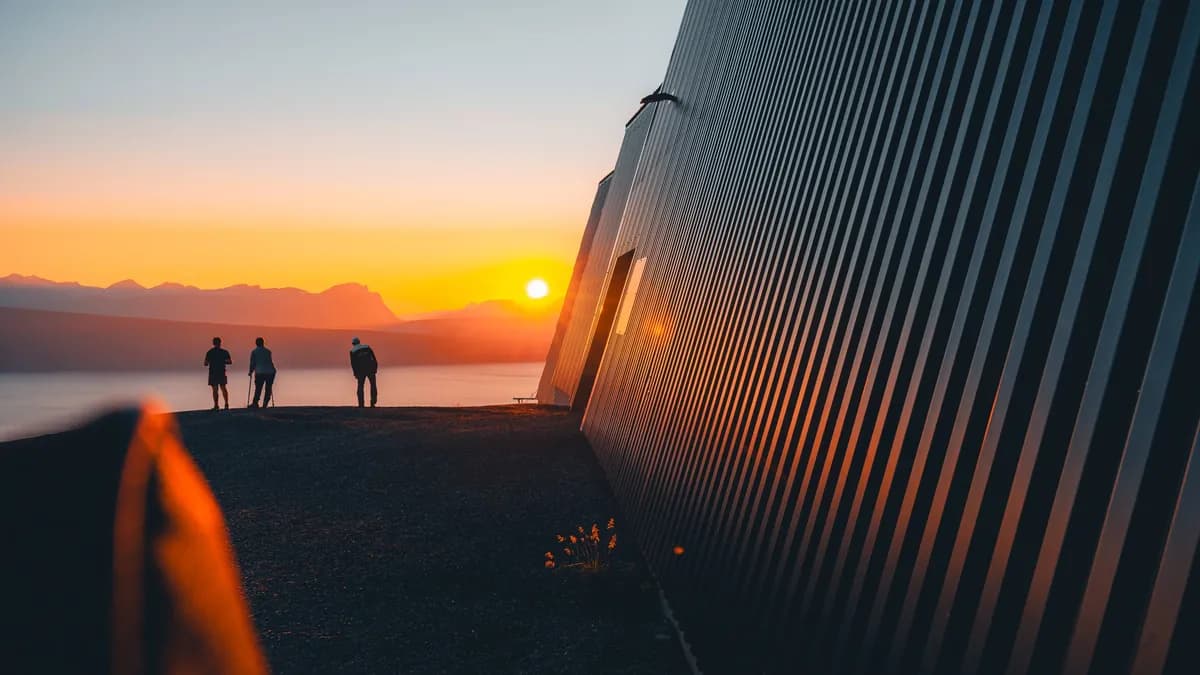 Three figures beside a ribbed cable-car station wall lit by the low orange sun.