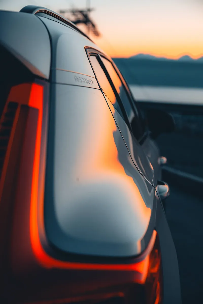 An orange sky reflected along a car beside a curved red tail light, with mountains beyond.
