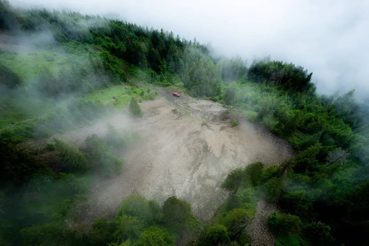 A tiny red car in a pale clearing surrounded by dense green forest and mist.