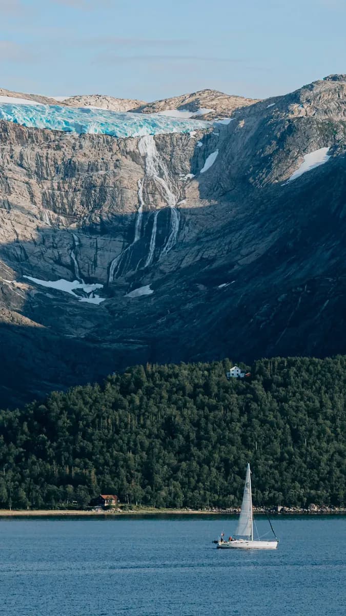 A small white sailboat below forest, exposed rock and a blue glacier.